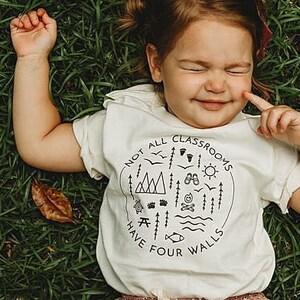Puede incluir: Un ni&ntilde;o peque&ntilde;o con una camiseta blanca con un gr&aacute;fico negro que dice "No todas las aulas tienen cuatro paredes". El gr&aacute;fico incluye una variedad de iconos al aire libre como monta&ntilde;as, &aacute;rboles, una fogata y un sol. El ni&ntilde;o est&aacute; acostado de espaldas en un campo de hierba.