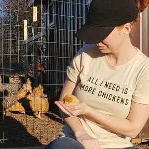 以下が含まれることがあります： ベージュのTシャツを着た女性が、小さな黄色いひよこを手に持っています。彼女は、鶏小屋の前でひざまずいており、背景にはいくつかの鶏がいます。Tシャツには「All I need is more chickens」とプリントされています。