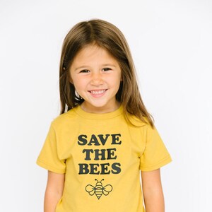Puede incluir: Una persona joven con una camiseta amarilla con el texto "SAVE THE BEES" y un gr&aacute;fico de abeja negro y amarillo. La persona lleva pantalones vaqueros azules.