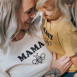 May include: A woman and a child wearing matching shirts with a bee graphic. The woman's shirt says "MAMA" and the child's shirt says "BABY".