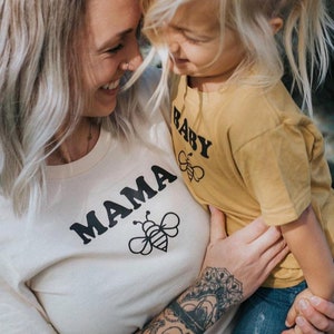 K&ouml;nnte beinhalten: Eine Frau und ein Kind tragen passende Shirts. Das Shirt der Frau sagt "MAMA" mit einer Bienengrafik. Das Shirt des Kindes sagt "BABY" mit einer Bienengrafik.