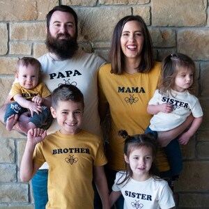May include: A family of five wearing matching shirts with bee designs. The shirts say "Papa", "Mama", "Brother", "Sister", and "Baby".