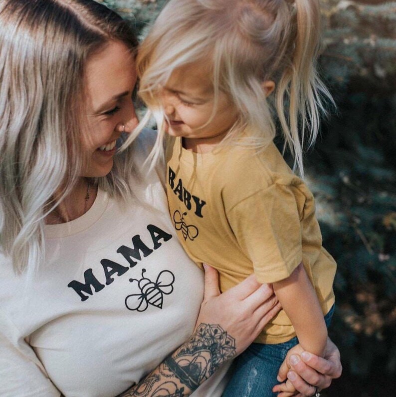 May include: A woman and a child wearing matching shirts with a bee graphic. The woman's shirt says "MAMA" and the child's shirt says "BABY".