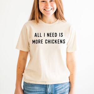 以下が含まれることがあります： クリーム色のTシャツを着た女性。Tシャツには「ALL I NEED IS MORE CHICKENS」という黒い文字がプリントされています。彼女は青いジーンズを履いています。