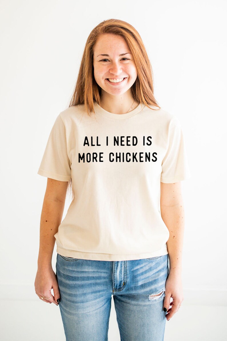 以下が含まれることがあります： クリーム色のTシャツを着た女性。Tシャツには「ALL I NEED IS MORE CHICKENS」という黒い文字がプリントされています。彼女は青いジーンズを履いています。