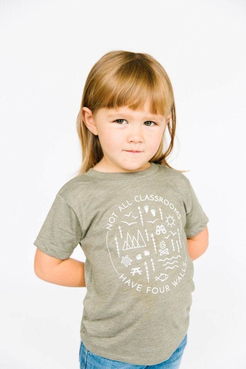 Puede incluir: Una persona joven lleva una camiseta verde con un gr&aacute;fico blanco que dice "No todas las aulas tienen cuatro paredes". El gr&aacute;fico incluye ilustraciones de monta&ntilde;as, &aacute;rboles, una fogata, un pez, una br&uacute;jula, una tienda de campa&ntilde;a y una mesa de picnic.