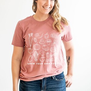 May include: A woman wearing a mauve t-shirt with a white graphic of various fruits and vegetables. The text "GROW YOUR OWN FOOD" is printed on the bottom of the shirt.