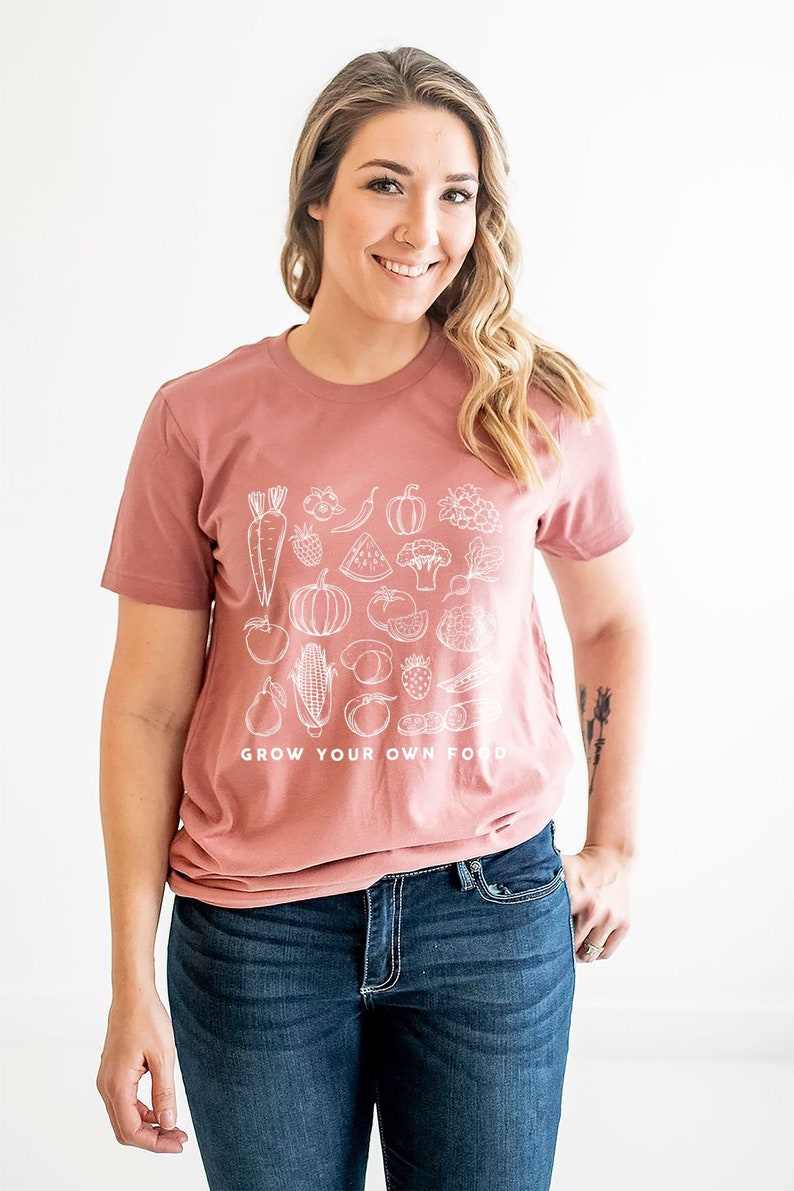 May include: A woman wearing a mauve t-shirt with a white graphic of various fruits and vegetables. The text "GROW YOUR OWN FOOD" is printed on the bottom of the shirt.