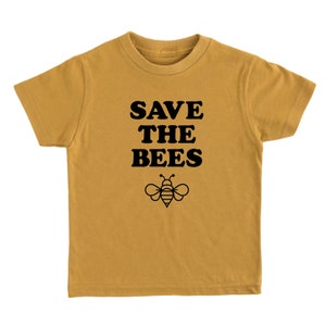 Puede incluir: Una camiseta de manga corta de color amarillo mostaza con el texto "SAVE THE BEES" en letras negras en bloque y una silueta negra de una abeja debajo del texto.