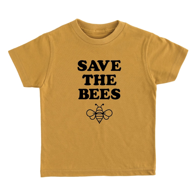 Puede incluir: Una camiseta de manga corta de color amarillo mostaza con el texto "SAVE THE BEES" en letras negras en bloque y una silueta negra de una abeja debajo del texto.