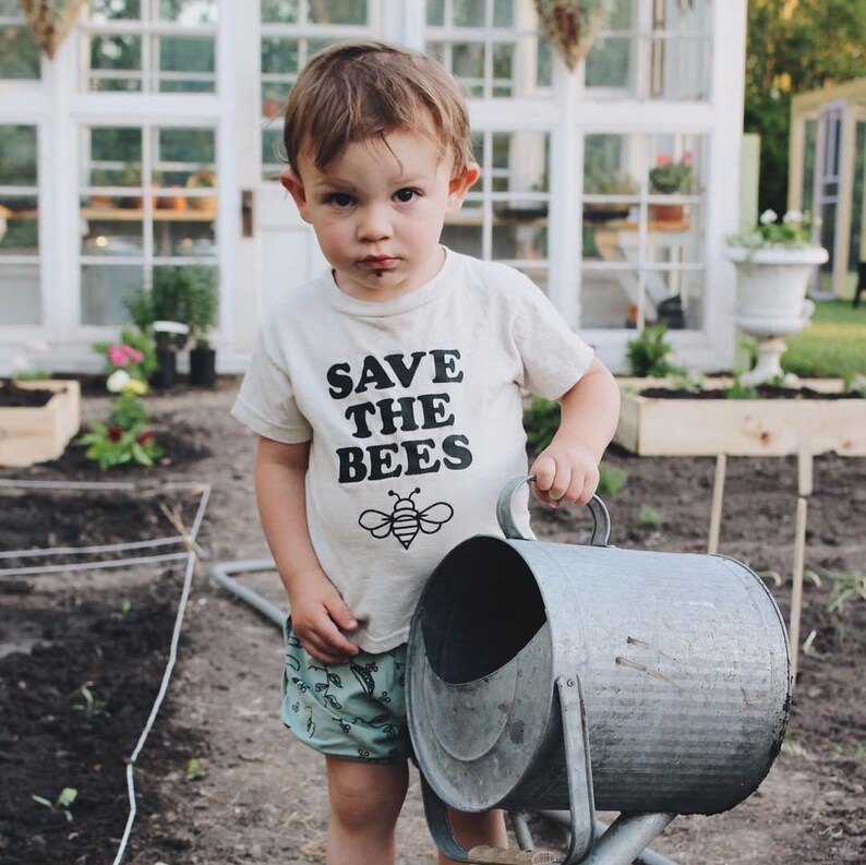 Puede incluir: Un ni&ntilde;o peque&ntilde;o con una camiseta blanca con el texto "Save the Bees" y un gr&aacute;fico de abeja. El ni&ntilde;o sostiene una regadera de metal en un jard&iacute;n.