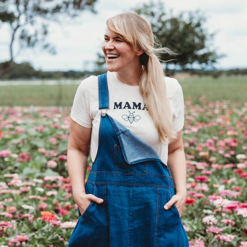 May include: A woman wearing a white t-shirt with the text "MAMA" and a bee graphic, and blue denim overalls, standing in a field of pink flowers.