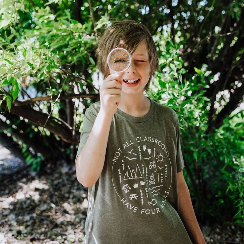Puede incluir: Una persona joven lleva una camiseta verde con un gr&aacute;fico blanco que dice "No todas las aulas tienen cuatro paredes". El gr&aacute;fico incluye un c&iacute;rculo con una cordillera, un sol, un p&aacute;jaro, un &aacute;rbol, una br&uacute;jula, un pez, una tortuga, una fogata y una huella. La persona sostiene una lupa frente a su ojo y sonr&iacute;e.