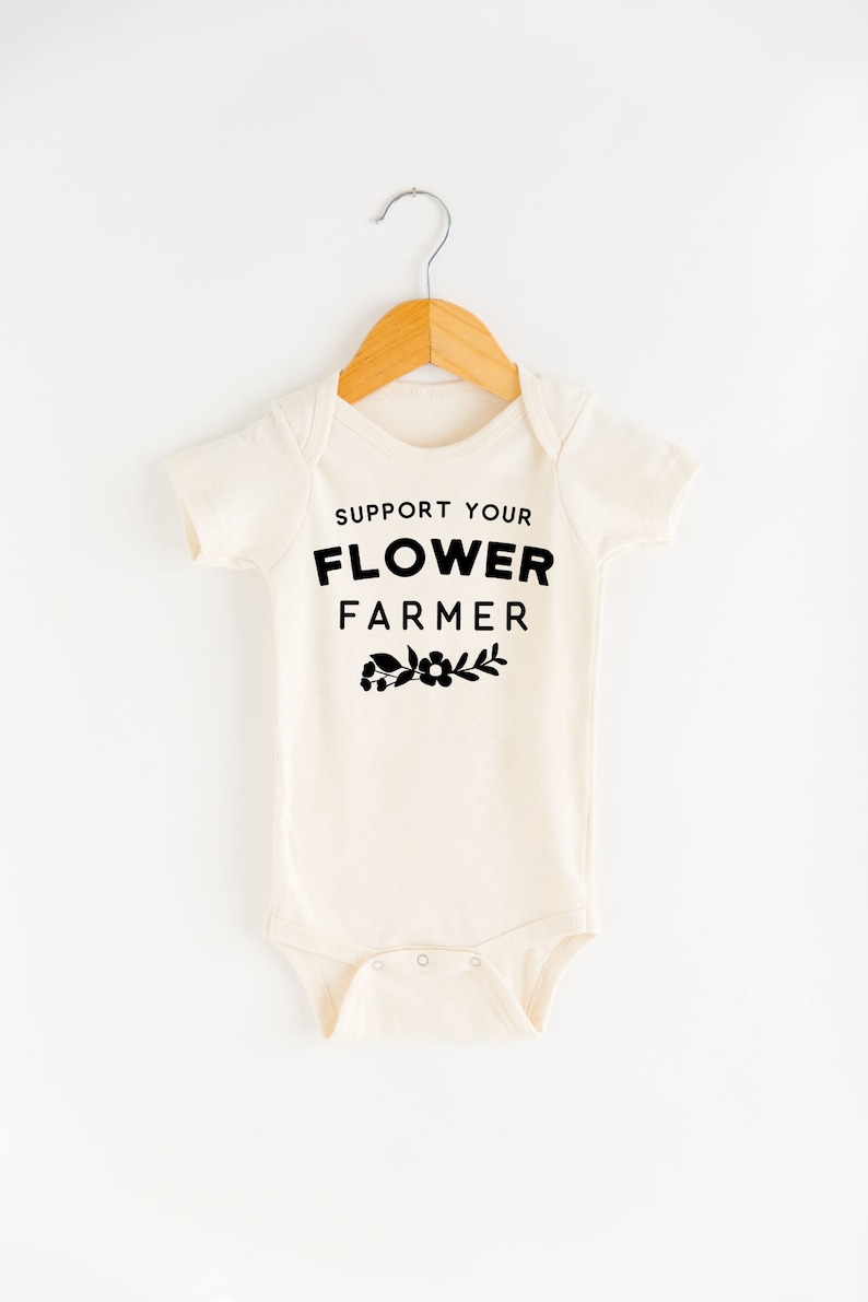 May include: A cream-colored baby onesie with short sleeves and black text that reads "Support Your Flower Farmer" with a small flower graphic below the text.