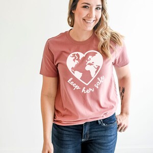 May include: A mauve t-shirt with a white graphic of the earth inside a heart. The text "keep her safe" is written below the graphic.