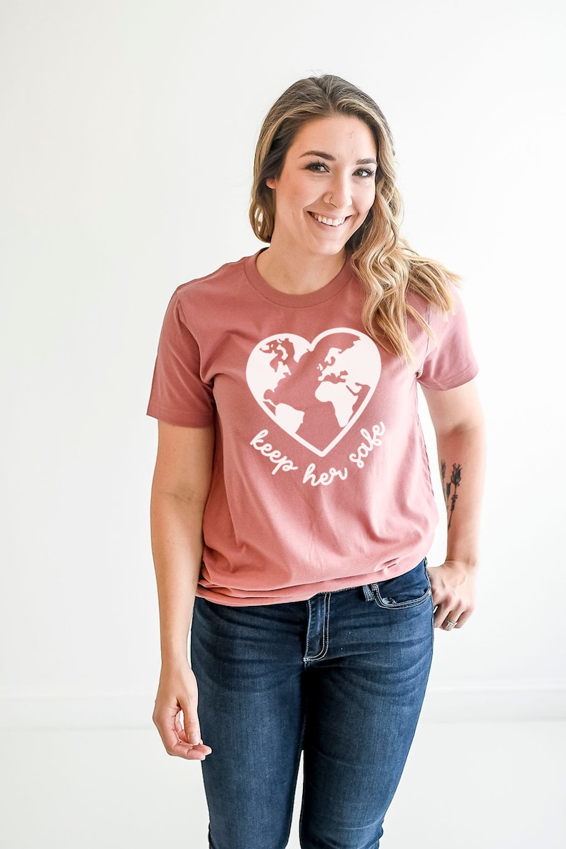 May include: A mauve t-shirt with a white graphic of the earth inside a heart. The text "keep her safe" is written below the graphic.