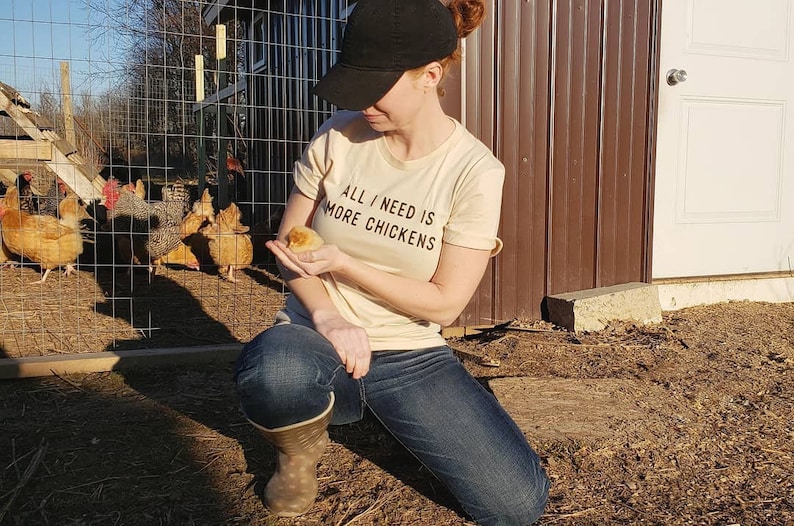 以下が含まれることがあります： ベージュのTシャツを着た女性が、小さな黄色いひよこを手に持っています。彼女は、鶏小屋の前でひざまずいており、背景にはいくつかの鶏がいます。Tシャツには「All I need is more chickens」とプリントされています。