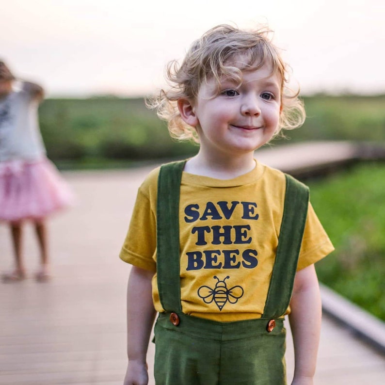 Puede incluir: Un ni&ntilde;o peque&ntilde;o con una camiseta amarilla con el texto "SAVE THE BEES" y un gr&aacute;fico de abeja. El ni&ntilde;o tambi&eacute;n lleva un mono verde con botones marrones.