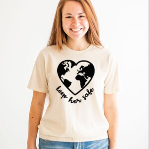 May include: A woman wearing a cream colored t-shirt with a black graphic of a heart-shaped earth and the text "keep her safe". She is wearing blue jeans.