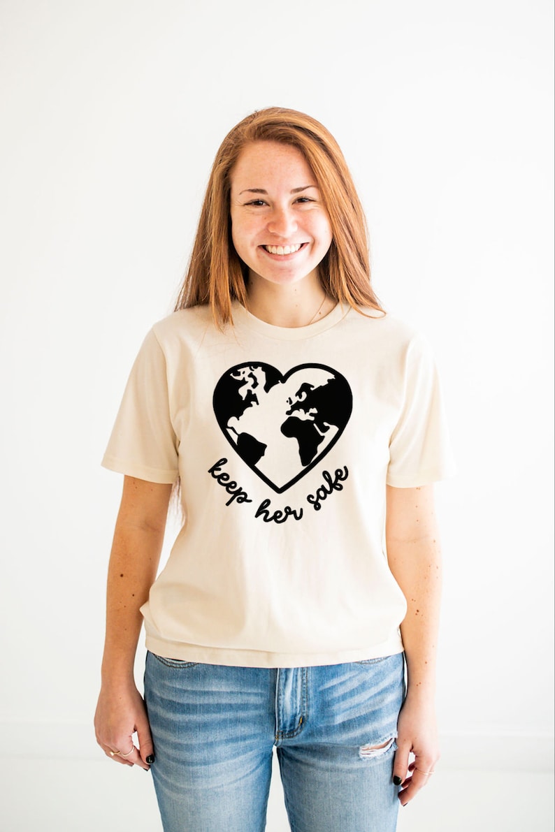May include: A woman wearing a cream colored t-shirt with a black graphic of a heart-shaped earth and the text "keep her safe". She is wearing blue jeans.
