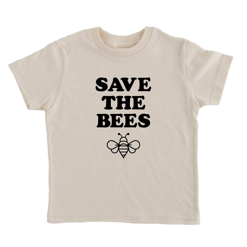 Puede incluir: Una camiseta de color crema con texto negro que dice "SAVE THE BEES" y una ilustraci&oacute;n de abeja negra.