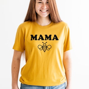 K&ouml;nnte beinhalten: Ein senfgelbes T-Shirt mit schwarzem Text, der "MAMA" lautet, und einer schwarzen Bienengrafik.