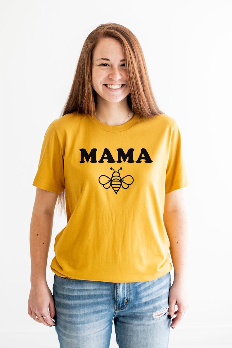 K&ouml;nnte beinhalten: Ein senfgelbes T-Shirt mit schwarzem Text, der "MAMA" lautet, und einer schwarzen Bienengrafik.