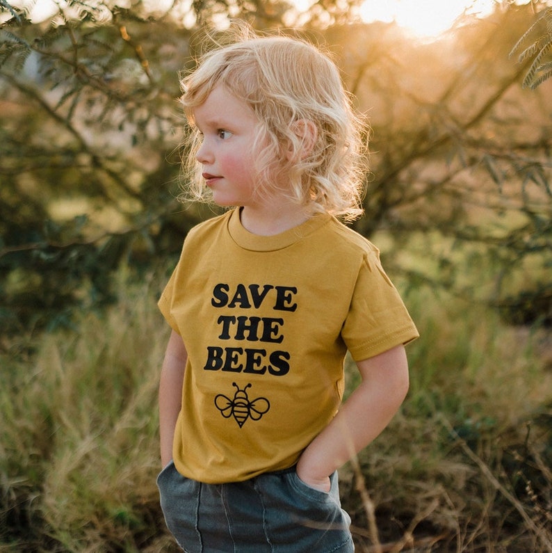 Puede incluir: Un ni&ntilde;o o ni&ntilde;a lleva una camiseta amarilla mostaza con el texto "SAVE THE BEES" y un peque&ntilde;o gr&aacute;fico de abeja. El ni&ntilde;o o ni&ntilde;a est&aacute; de pie en un campo de hierba alta.