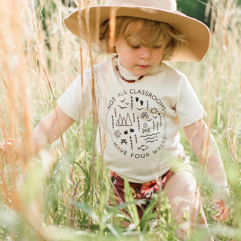 Puede incluir: Un ni&ntilde;o con una camiseta blanca con un gr&aacute;fico negro que dice "No todas las aulas tienen cuatro paredes" y un sombrero marr&oacute;n est&aacute; de pie en un campo de hierba alta.
