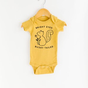 May include: A mustard yellow baby onesie with a black and white illustration of a squirrel holding a flower. The text "BRIGHT EYED BUSHY TAILED" is printed on the onesie.