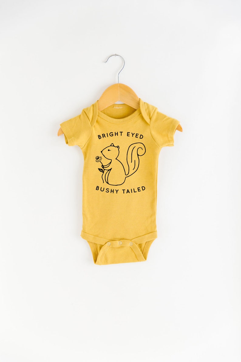 May include: A mustard yellow baby onesie with a black and white illustration of a squirrel holding a flower. The text "BRIGHT EYED BUSHY TAILED" is printed on the onesie.