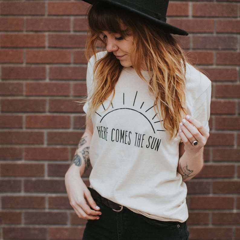 K&ouml;nnte beinhalten: Ein wei&szlig;es T-Shirt mit einer schwarzen Grafik einer Sonne und dem Text "Here Comes The Sun".