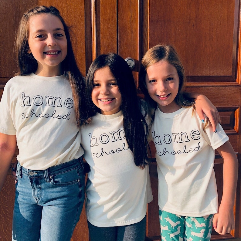 May include: Three girls wearing white t-shirts with the text "home schooled" printed on them. The girls are standing in front of a wooden door.
