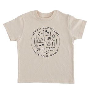 Puede incluir: Una camiseta beige de manga corta con un estampado gr&aacute;fico negro en forma de c&iacute;rculo con el texto "Not all classrooms have four walls" y varios iconos de exteriores, incluyendo monta&ntilde;as, &aacute;rboles, un sol, un pez, una br&uacute;jula y huellas.