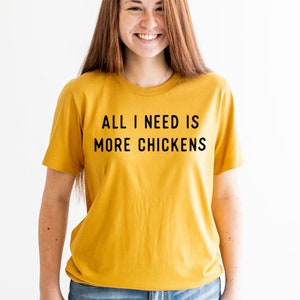 以下が含まれることがあります： 黒い文字で「All I need is more chickens」と書かれたマスタードイエローのTシャツを着た女性。彼女は笑顔で、青いジーンズを履いています。
