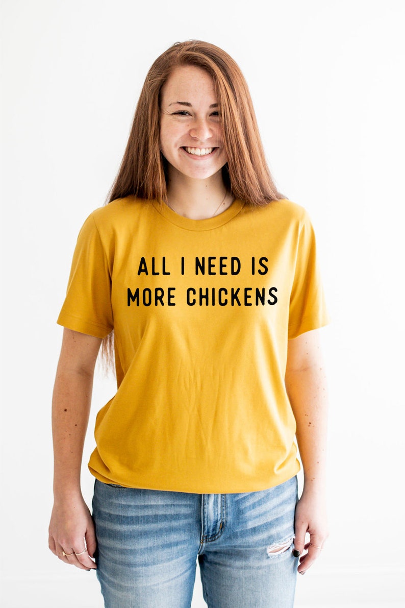 以下が含まれることがあります： 黒い文字で「All I need is more chickens」と書かれたマスタードイエローのTシャツを着た女性。彼女は笑顔で、青いジーンズを履いています。