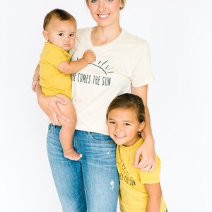 K&ouml;nnte beinhalten: Eine Frau tr&auml;gt ein cremefarbenes T-Shirt mit dem Text "Here comes the sun" und blaue Jeans und h&auml;lt ein Baby in einem gelben Strampler. Ein junges M&auml;dchen tr&auml;gt ein gelbes T-Shirt mit dem Text "Here comes the sun" und blaue Jeans-Shorts und steht neben der Frau.