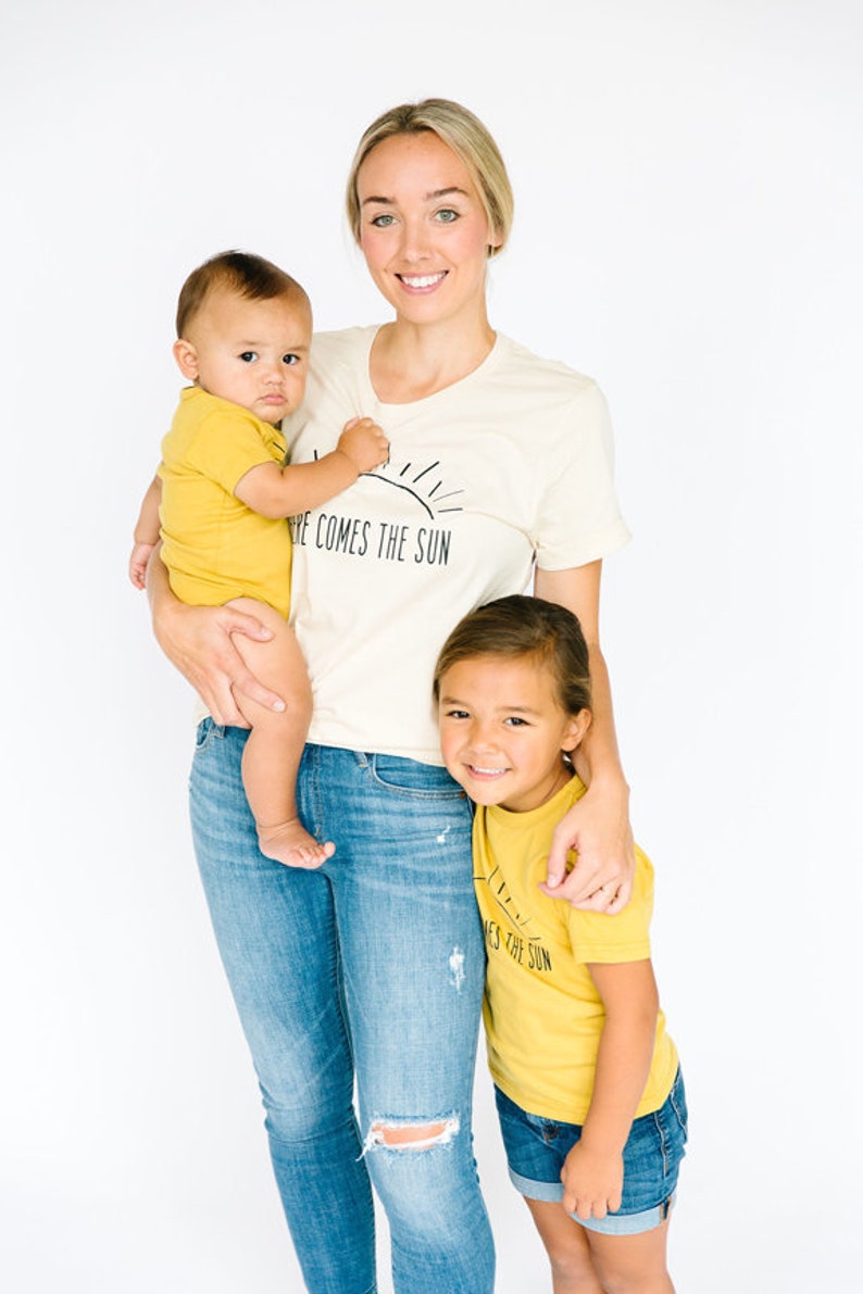 K&ouml;nnte beinhalten: Eine Frau tr&auml;gt ein cremefarbenes T-Shirt mit dem Text "Here comes the sun" und blaue Jeans und h&auml;lt ein Baby in einem gelben Strampler. Ein junges M&auml;dchen tr&auml;gt ein gelbes T-Shirt mit dem Text "Here comes the sun" und blaue Jeans-Shorts und steht neben der Frau.
