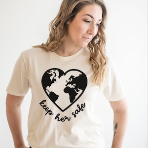 May include: A cream colored t-shirt with a black graphic of the earth in a heart shape. The text "keep her safe" is written below the graphic.
