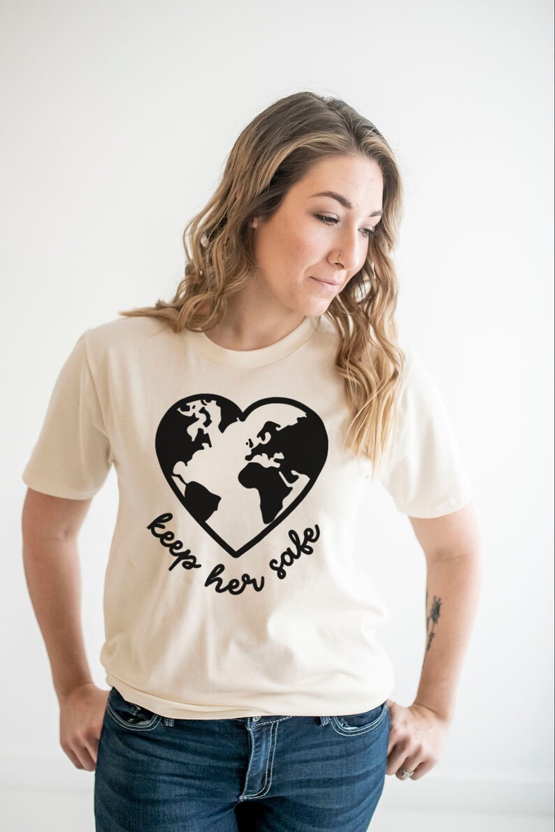 May include: A cream colored t-shirt with a black graphic of the earth in a heart shape. The text "keep her safe" is written below the graphic.