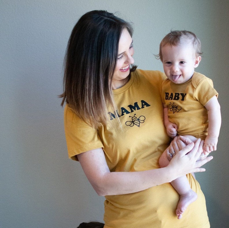 K&ouml;nnte beinhalten: Eine Frau und ein Baby tragen passende T-Shirts in Senfgelb. Das Shirt der Frau tr&auml;gt den Schriftzug "Mama" mit einer Bienengrafik. Das Shirt des Babys tr&auml;gt den Schriftzug "Baby" mit einer Bienengrafik.