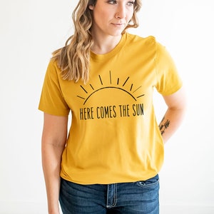 K&ouml;nnte beinhalten: Ein senfgelbes T-Shirt mit einer schwarzen Grafik einer Sonne und dem Text "Here Comes The Sun".