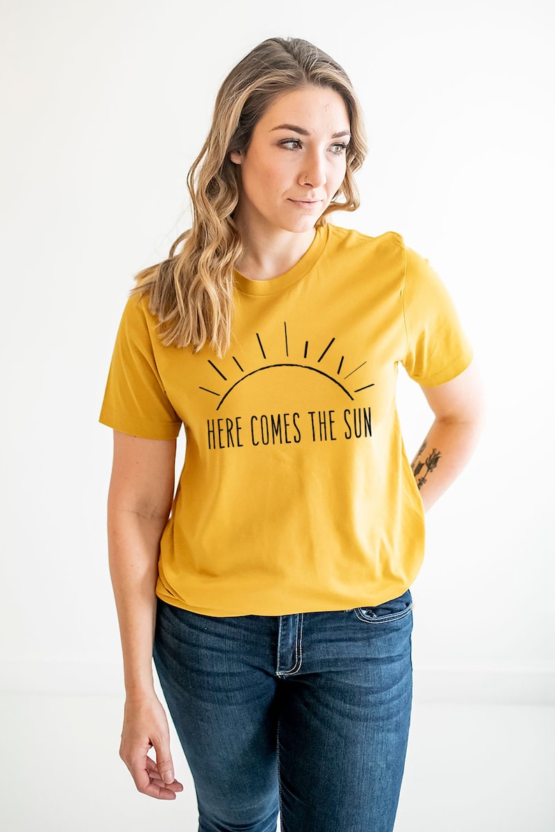 K&ouml;nnte beinhalten: Ein senfgelbes T-Shirt mit einer schwarzen Grafik einer Sonne und dem Text "Here Comes The Sun".