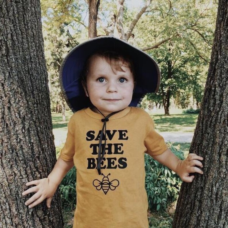 Puede incluir: Un ni&ntilde;o peque&ntilde;o con una camiseta amarilla con el texto "SAVE THE BEES" y un gr&aacute;fico de una abeja. El ni&ntilde;o lleva un sombrero de sol azul y est&aacute; de pie entre dos &aacute;rboles.
