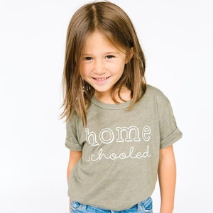 May include: A young girl wearing a green t-shirt with the text "home schooled" in white. She is smiling and wearing blue jeans.