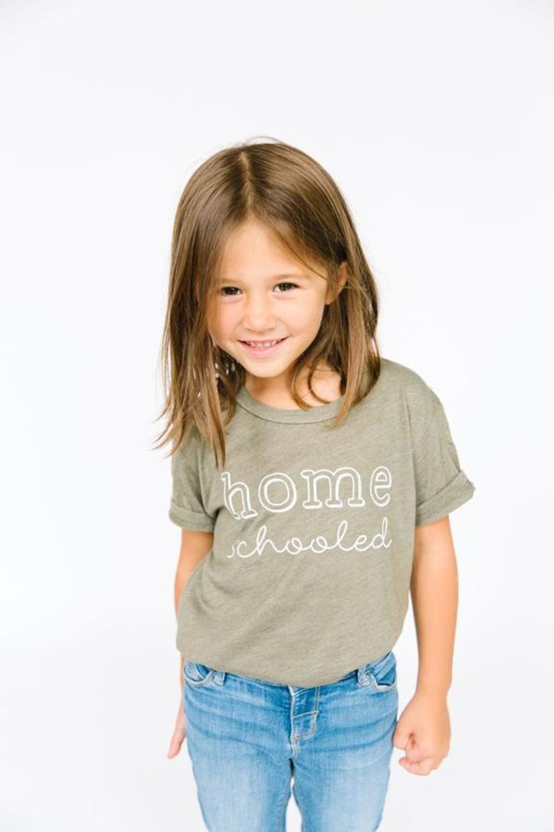 May include: A young girl wearing a green t-shirt with the text "home schooled" in white. She is smiling and wearing blue jeans.