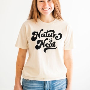 May include: A woman wearing a cream colored t-shirt with the text "Nature is Neat" in black script. She is wearing blue jeans.