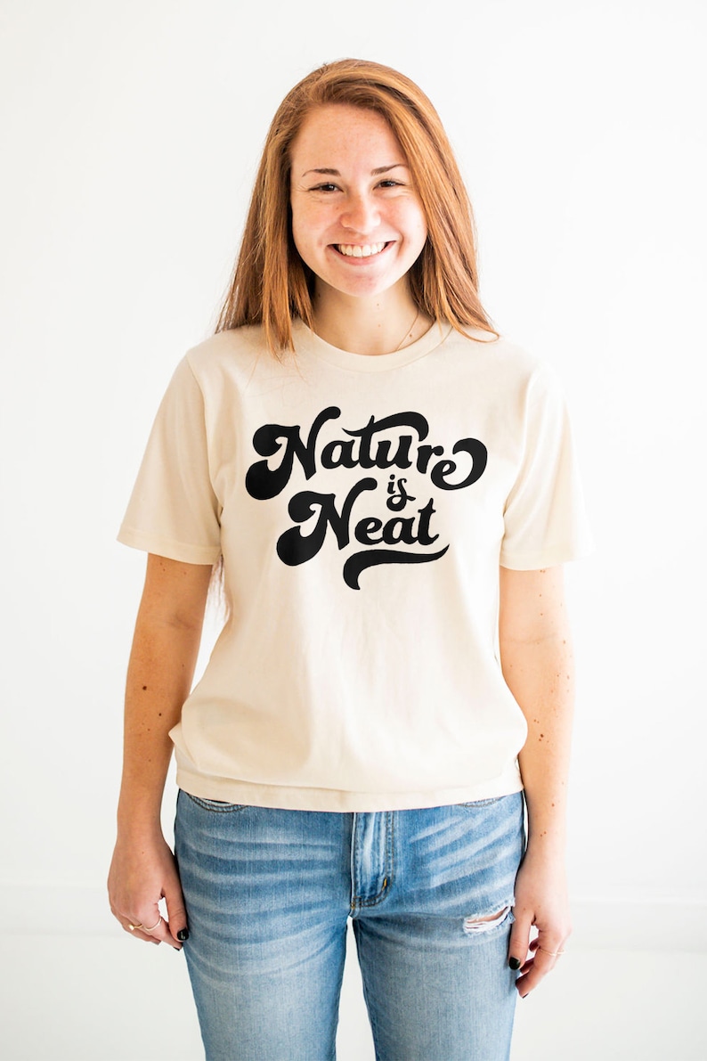 May include: A woman wearing a cream colored t-shirt with the text "Nature is Neat" in black script. She is wearing blue jeans.