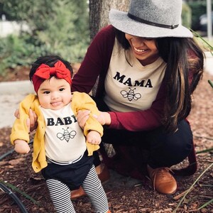 May include: A mother and child wearing matching shirts. The mother's shirt says "Mama" with a bee graphic. The child's shirt says "Baby" with a bee graphic. The child is wearing a yellow cardigan and black and white striped leggings.