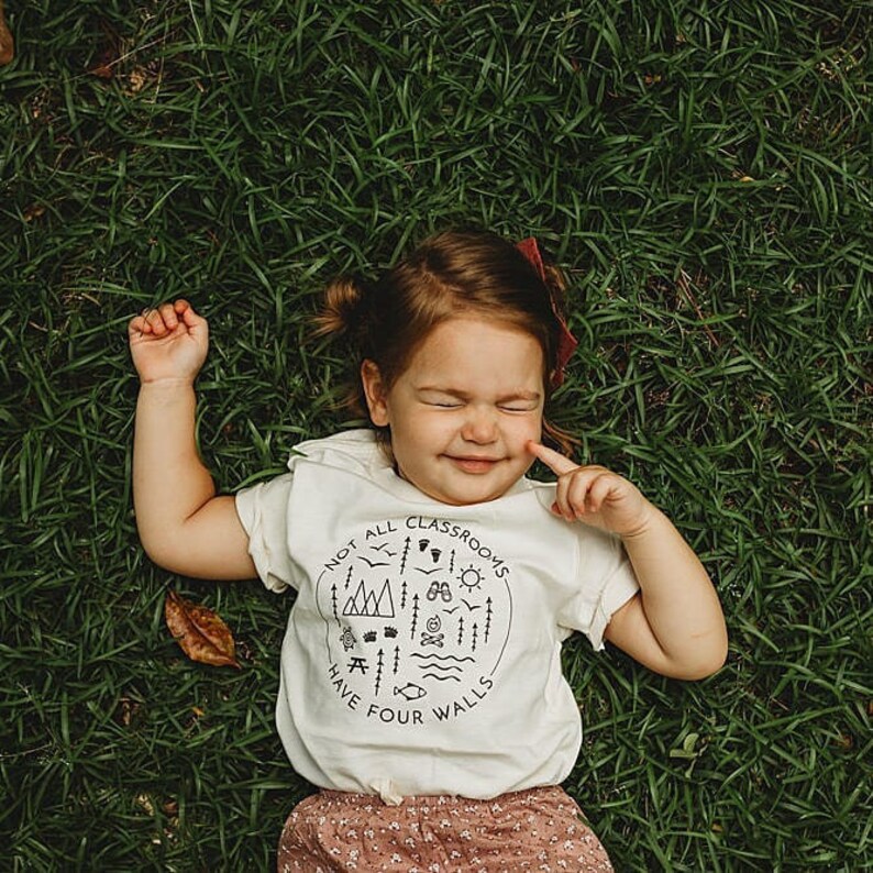 Puede incluir: Un ni&ntilde;o peque&ntilde;o con una camiseta blanca con un gr&aacute;fico negro que dice "No todas las aulas tienen cuatro paredes". El gr&aacute;fico incluye una variedad de iconos al aire libre como monta&ntilde;as, &aacute;rboles, una fogata y un sol. El ni&ntilde;o est&aacute; acostado de espaldas en un campo de hierba.